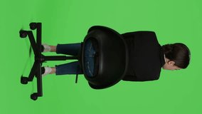 Vertical video: Back view of caucasian businesswoman wearing suit and sitting on chair, waiting in preparation over full body green screen backdrop. Young adult working as corporate employee being