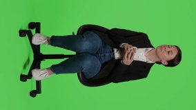 Vertical video: Front view of casual company worker drinking cup of coffee over full body greenscreen, sitting on chair in studio. Young employee wearing office suit and enjoying caffeine beverage