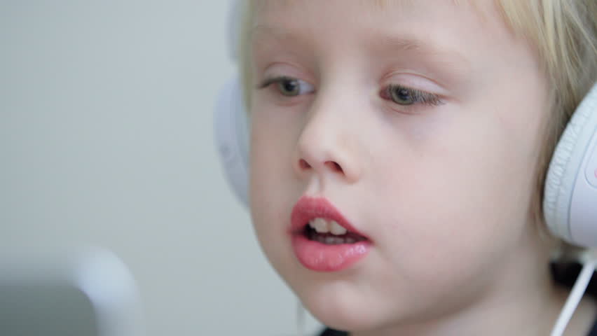 Close portrait of sweet caucasian boy with blue eyes and in headphones looking at something or someone and repeating words. Online speech lesson for a 5 years old boy