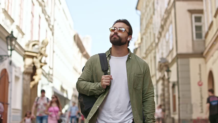 Front view of male tourist traveling, admiring, watching architecture, buildings, smiling. Handsome man with beard carrying backpack, listening to music. Concept of tourism.