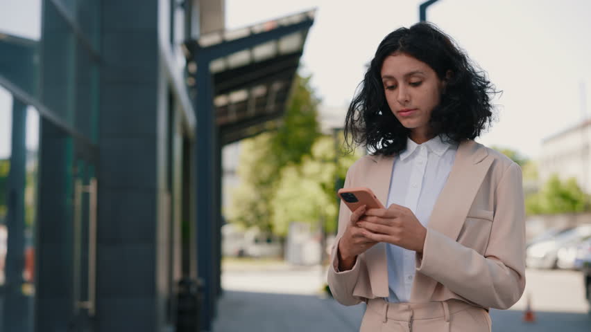 Beautiful curly smiling young businesswoman in formal outfit standing near the business centre using smartphone. People and technology business concept | Shutterstock HD Video #1098192185