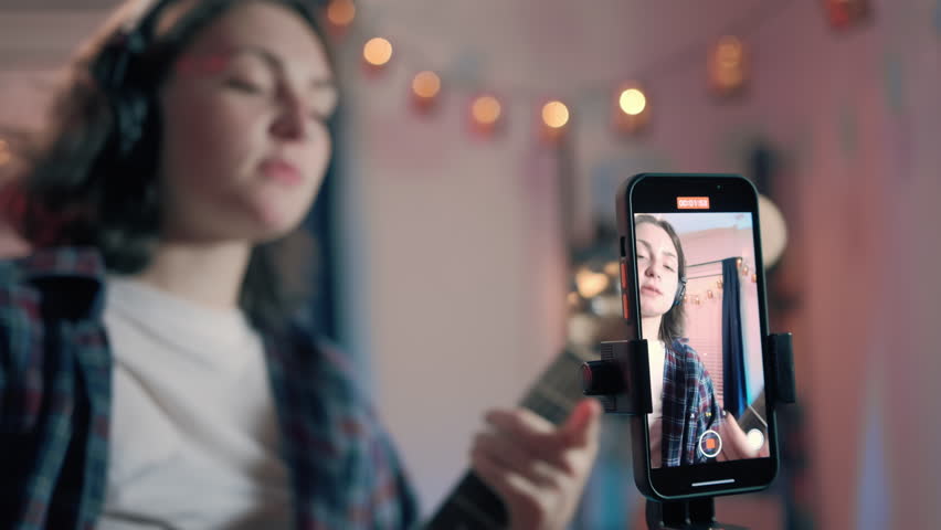 Portrait Art Woman Bloggers, Online Lesson Recording, Playing Guitar. Cheerful woman blogger playing guitar while sitting using mobile phone. Focus of smartphone