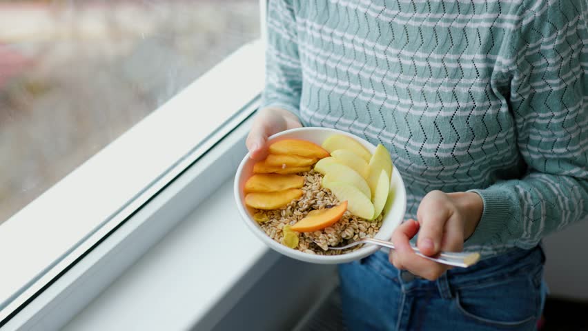 Close up of hands of woman holding homemade granola in a plate with fruits, grain, oatmeal and other natural ingredients. Focus on a bowl with healthy breakfast, carbohydrates and coarse fibers.
