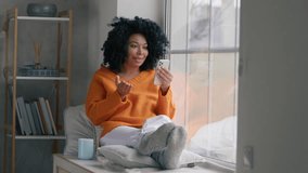Black smiling woman using app on cellphone at home. Beautiful relaxed girl chatting on mobile phone looking at mobile camera 4K. Cheerful young African woman using smartphone while sitting at window