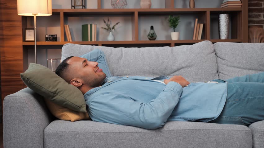 Happy young man laying on sofa, relaxed african american guy resting napping on sofa with eyes closed at home, healthy guy breathing fresh air lounge on couch hands behind head enjoy stress free. | Shutterstock HD Video #1098217781