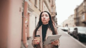 Beautiful young caucasian business woman walks at city street and uses tablet to type text or receive information, holds coffee. Online communication or business work problems in remote freelance.