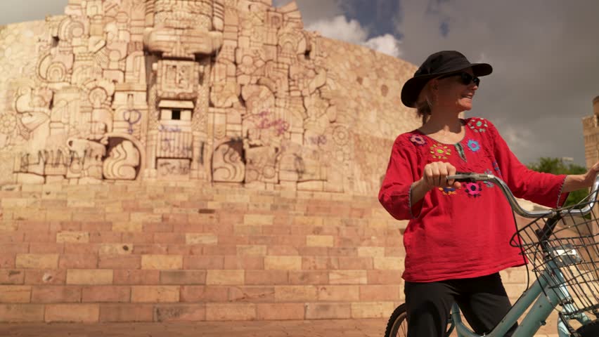 Portrait of smiling, happy, fun mature elderly woman in ethnic clothing having fun biking on vacation in front of the Monumento a la Patria monument in Merida, Yucatan, Mexico.