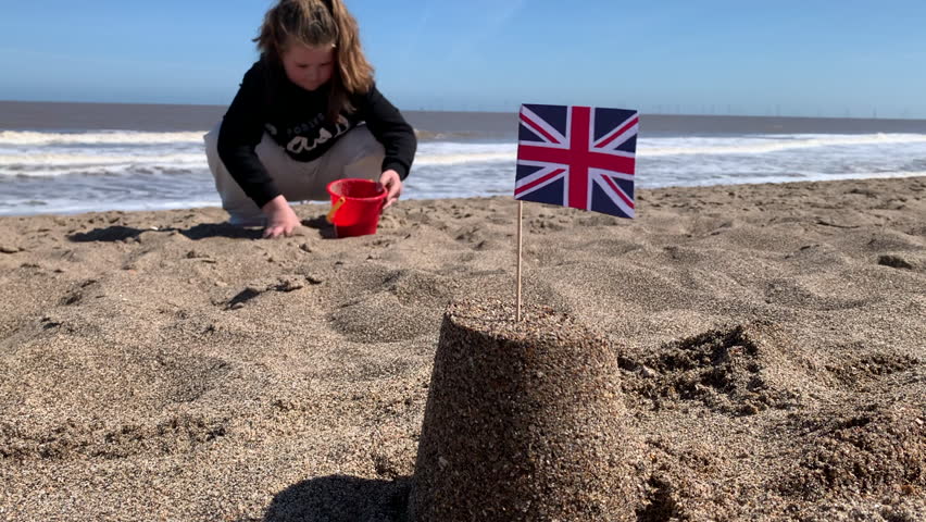 A girl fillin a bucket with sand to build sandcastles on a British beach. The UK union jack is on top of the sand castles with the sea waves rolling in on a staycation holiday on the coast in England. | Shutterstock HD Video #1098232265