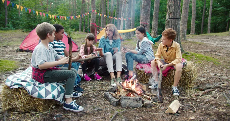 Happy caucasian woman sitting at tent in forest with her pupils. Children and their teacher resting in wood and having pleasant conversation at camping. Nature and travel concept | Shutterstock HD Video #1098234229
