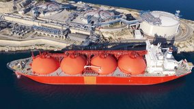 Aerial drone pullback video of LNG (Liquified Natural Gas) tanker anchored in small gas terminal island with tanks for storage