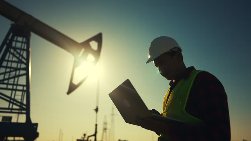 Oil business. a worker works next to an oil pump holding lifestyle a laptop. industry business oil and gas concept. engineer studying the level of oil production on a laptop silhouette at sunset