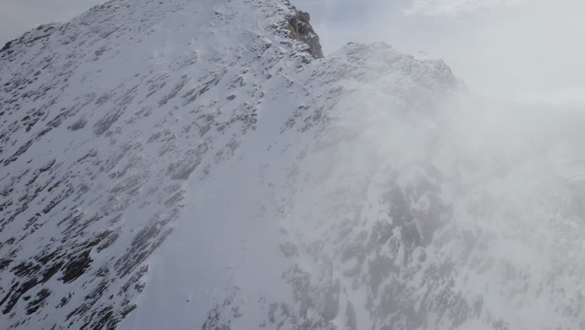 Aerial orbit shot of flight along rocky and snowy mountain wall in clouds of sky