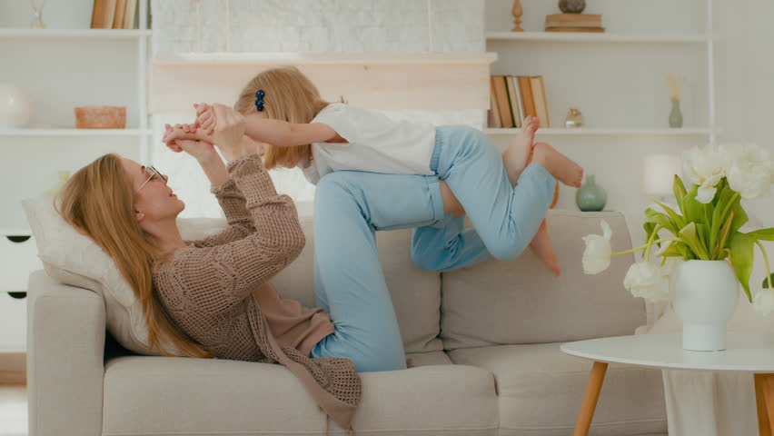 Caucasian mother in glasses strong mom lying on cozy couch on back holding little daughter in air on legs bent in knees play airplane flying game child girl preschooler kid baby imitate flight plane Royalty-Free Stock Footage #1098245327