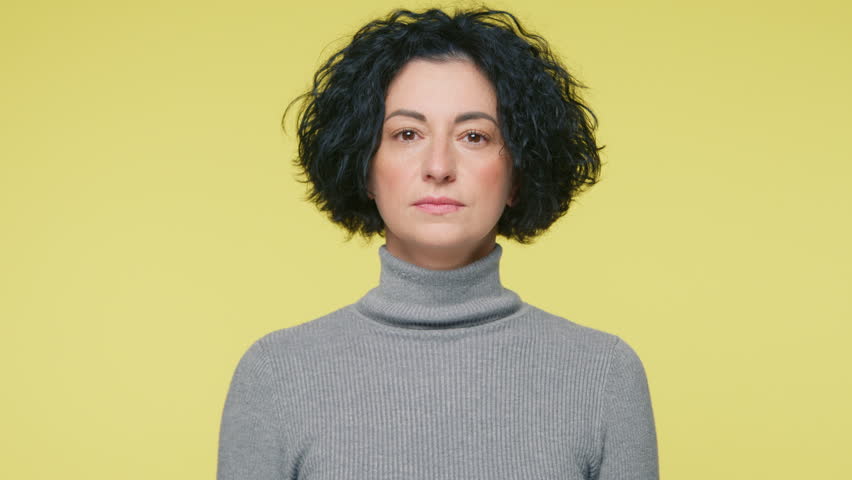 Closeup portrait of serious adult female looking at camera threatening cutting throat with killing gesture of revenge aggression. Calm angry mature lady show sign cut hand neck on yellow background