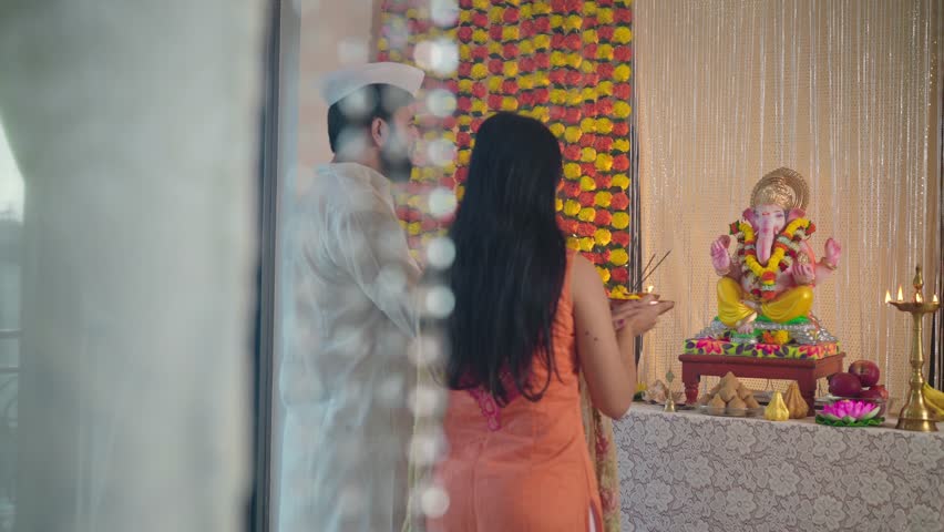 Happy ethnic Hindu Indian young married couple offering a prayer or worshiping the Ganapati idol decorated with marigold flowers during the Ganesha Chaturthi festival or celebration in an indoor house Royalty-Free Stock Footage #1098250299