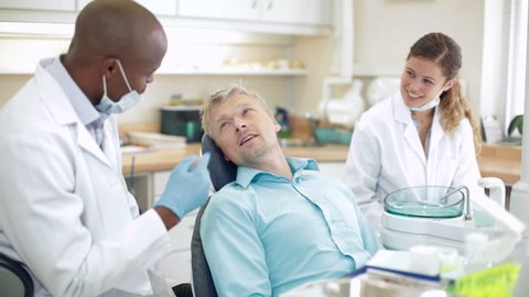 Scene in dentist office where dentist discusses treatment with smiling male patient who seems relaxed and happy. The smiling dental nurse looks on.