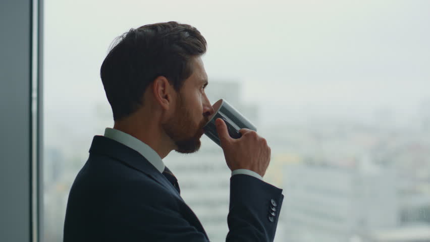 Rich company worker drinking hot beverage standing at office window close up. Attractive bearded executive manager creating new business ideas feeling inspiration. Entrepreneur anticipating success. | Shutterstock HD Video #1098253669