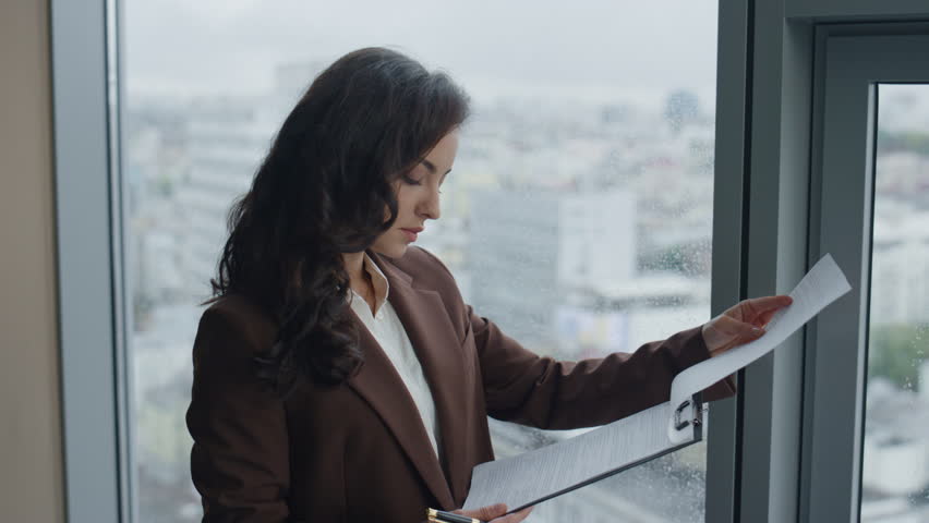 Attractive woman executive manager satisfied work documents standing alone at office window close up. Smiling beautiful brunette entrepreneur reading company contract profitful deal successful sales. Royalty-Free Stock Footage #1098253737