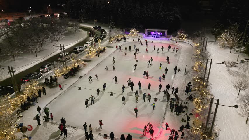 Families skating on outdoor ice rink surrounded by Christmas lights at night with DJ booth and lights set up behind