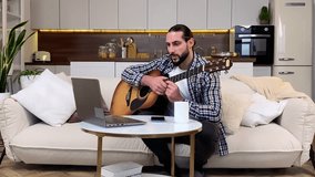 Handsome middle aged bearded man watching video tutorial to learn playing guitar by himself indoors. Happy male person taking online lessons via Internet using laptop at home. Domestic hoppy concept