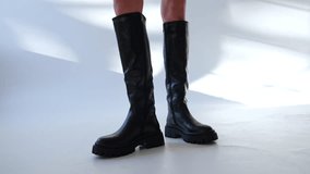 High black leather boots on plain tractor soles on the bare female legs. Model showing the footwear in light studio.