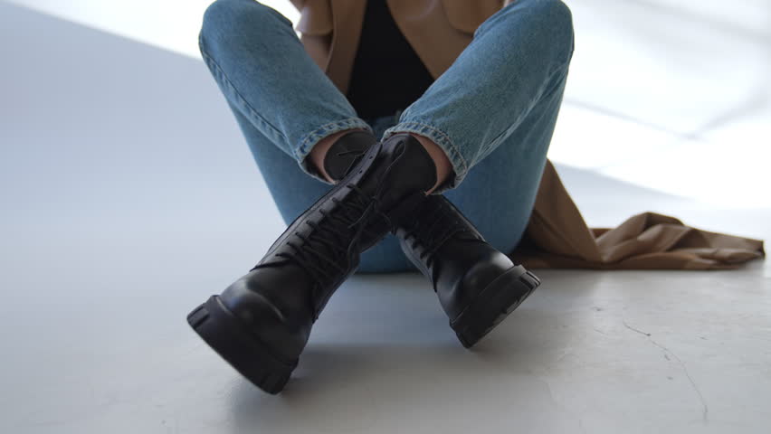 Female sits on the floor her stylish black boots near camera. Close up. Woman in jeans and coat showing her modern footwear.