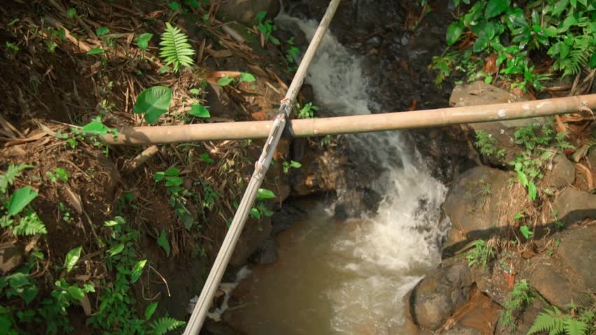 A high-angle, close-up shot of a small, fast-flowing waterfall or irrigation stream cascading over rocks. A simple bamboo pipe crosses the water, surrounded by green ferns and plants.