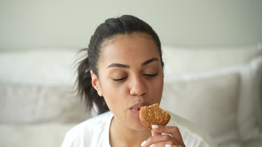 Pretty African American girl in casual eating cookie smiling looking at camera enjoys Sunday at home. Adorable Brazilian young woman likes bakery offering to eat together. Happy people at home Royalty-Free Stock Footage #1098272929