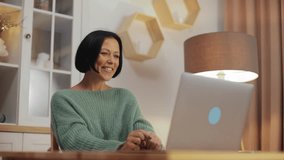 Portrait of a charming middle-aged woman sitting at a table in a cozy living room and talking on a laptop during a video chat meeting. Freelance or remote work, video call with friends or relatives