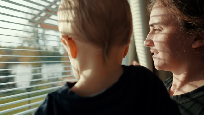 Woman holds toddler in arms looking at lake through blinds on window on sunny morning. Mother and son enjoy weekend in cottage house closeup