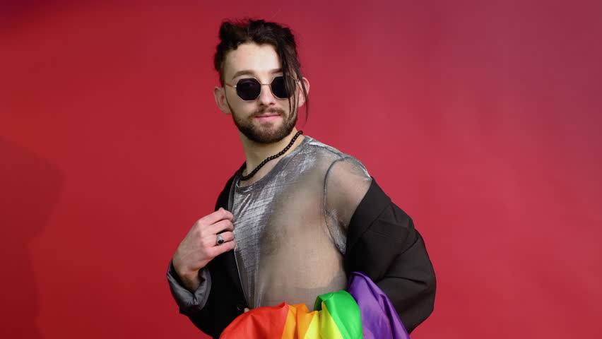Gay man dressed in mesh t-shirt and sunglasses on red background with a multicolored rainbow flag. Concept diversity, transsexual, and freedom