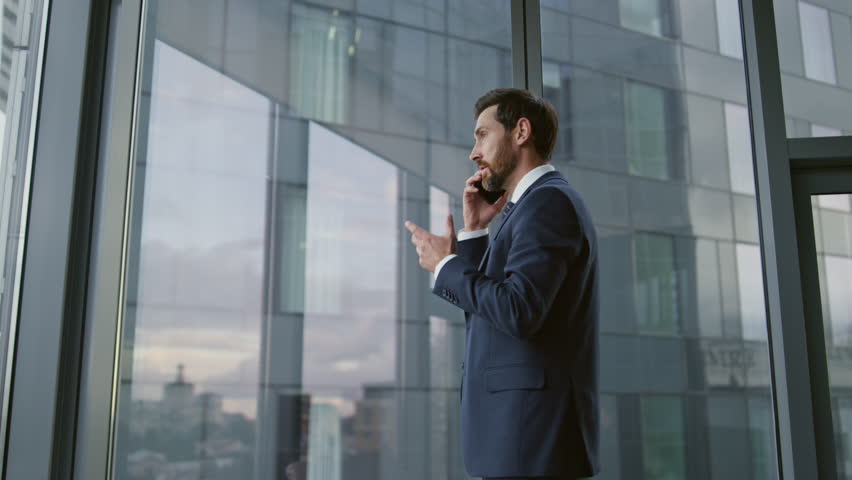 Worried ceo calling phone standing near office window close up. Confident bearded executive manager have serious telephone conversation feeling stress. Busy nervous business man finish smartphone talk Royalty-Free Stock Footage #1098288637