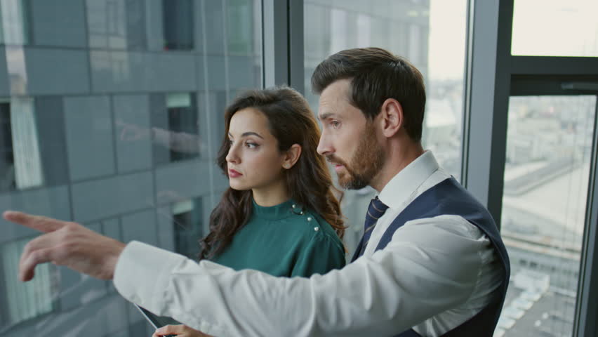 Busy confident partners discussing business project standing near panoramic window looking on city view close up. Serious colleagues working together in modern office. Couple talking about strategy.