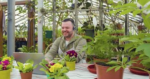 A young greenhouse worker during an online video call - he sitting in front of laptop and telling customers about the grown flowers to the camera and actively gesticulates. 4k footage.