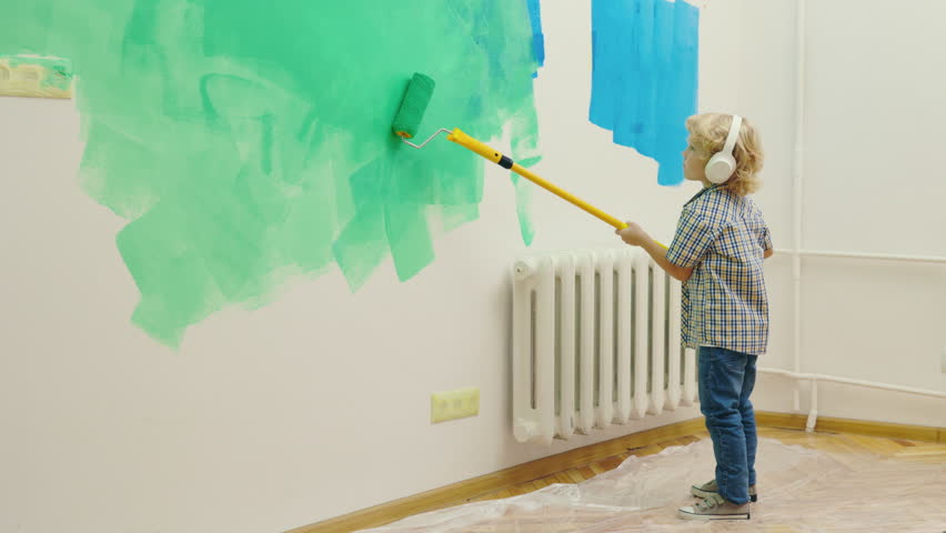 Child painting wall with paint roller while listening to music. Adorable small boy paints with equipment. Kid helping parent with house renovation. Home repair Royalty-Free Stock Footage #1098304233