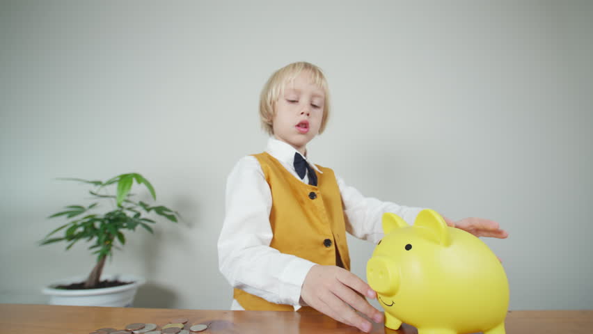 Cute caucasian boy in yellow suit and black tie-bow puts a few coins in big yellow money box. 5 years old boy counts and saves money in funny piggybank
