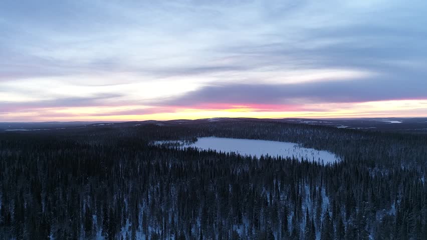 A small lake surrounded by dense snowy forests in winter in Lapland, Finland during sunset, drone