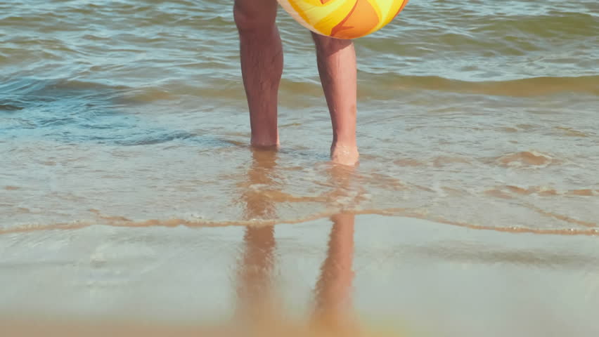 Tilt up of young bearded man in white shirt and yellow shorts holding inflatable pineapple shaped mattress while standing in water and looking at camera