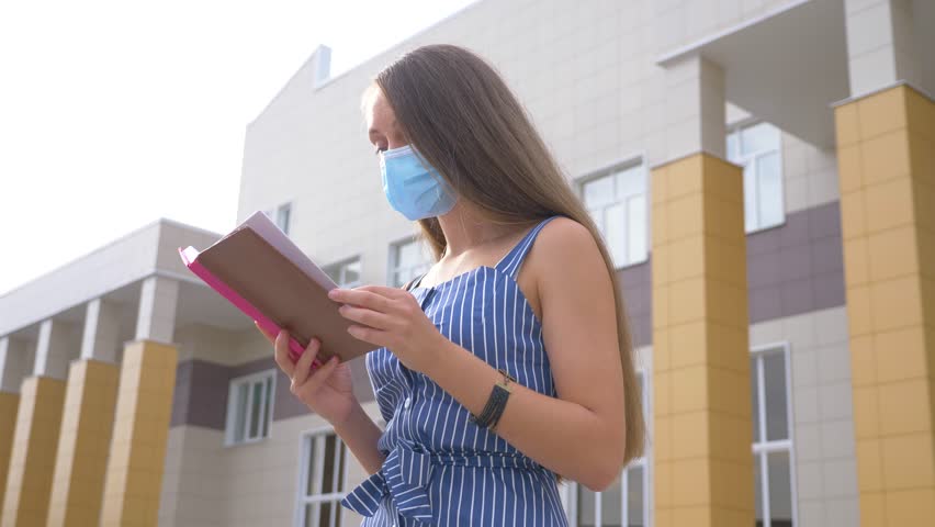 A girl reads a book in a Park with a medical mask on her face. A student is preparing for an exam remotely during the Covid-19 pandemic. a protective mask on the girl