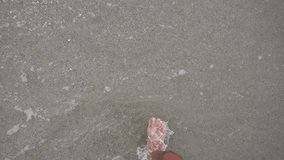 A top view of men's bare feet walking on sandy beach with small waves