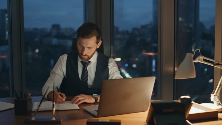 Overworked office manager typing on laptop sitting at company desk late night having overtime. Focused bearded businessman working feeling tired. Corporate employee looking on watch busy of paperwork. Royalty-Free Stock Footage #1098321021