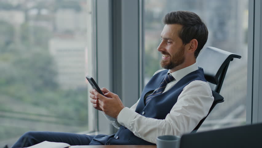 Satisfied entrepreneur watching phone receiving sms sitting at office in elegant suit close up. Confident bearded business man reading good news on smartphone screen. Manager taking break from work. | Shutterstock HD Video #1098321061