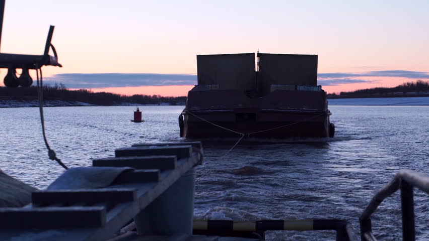 Barge in tow of fishing boat. Clip. Boat carries barge with cargo in cloudy twilight. Boat and barge float on river on winter morning
