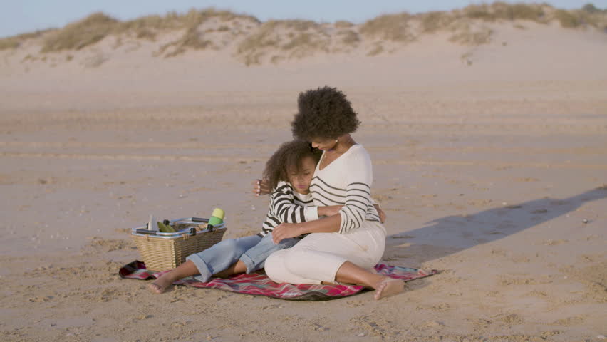 Happy mum and daughter having picnic on the beach in the evening. Pretty Black woman hugging her child sitting on blanket on sandy seashore, showing affection and smiling. Leisure, motherhood concept. | Shutterstock HD Video #1098325735