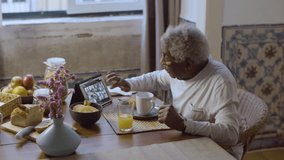 Elderly Black man having video chat at breakfast at home. Senior man enjoying orange juice and cupcakes, talking with friends on digital tablet, sharing news. Modern technology, retirement concept.