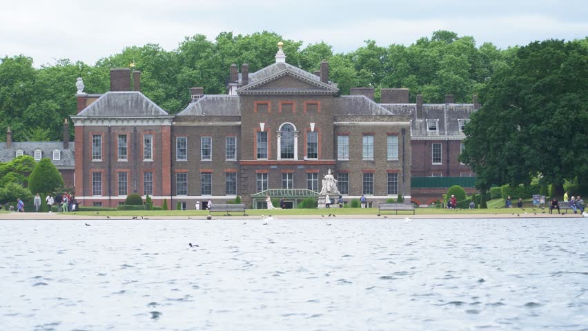 London, England - 27 May 2022: A beautiful historic royal London palace with a white statue in front overlooking a park with a large decorative pond with people walking past. 