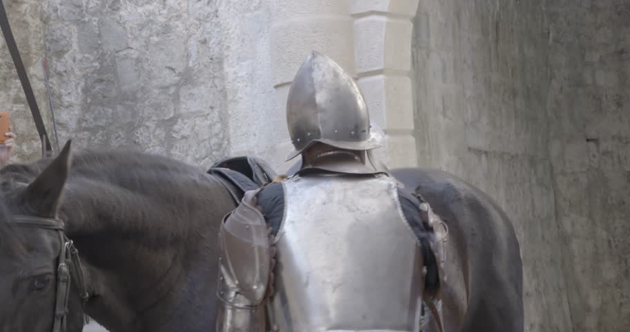 A knight in metal armor saddles a horse in front of the castle walls
