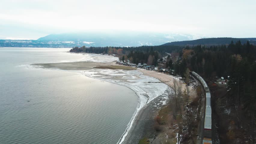 During the winter months near Salmon Arm, British Columbia, Canada, there is a train traveling along the tracks on a railroad next to Shuswap Lake, in a Cinematic Reverse Reveal shot.Canada.