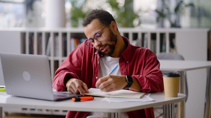 Successful young African American man, university graduate student, freelance entrepreneur, teacher talking on mobile phone, taking notes in notebook while working remotely on laptop in school library Royalty-Free Stock Footage #1098332251