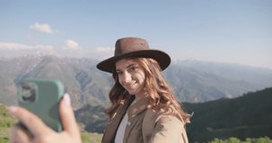 Beautiful brown-haired woman blogger in safari outfit takes selfie on smartphone against backdrop of mountains. Young caucasian woman with pale pink manicure takes selfie on smartphone for travel blog